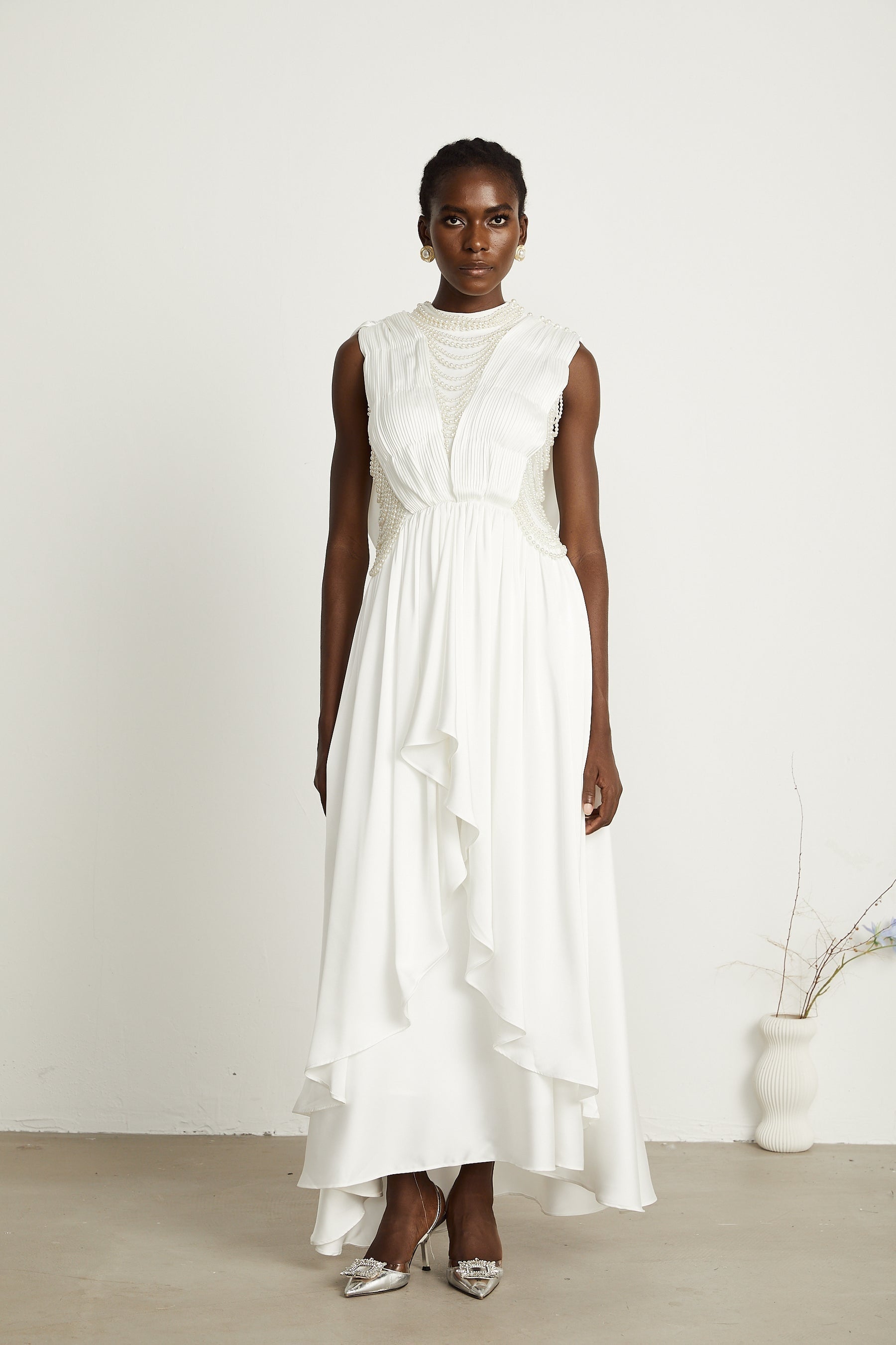 A long white maxi dress with pleated fabric and faux pearl embellishments worn by a model standing in a neutral setting