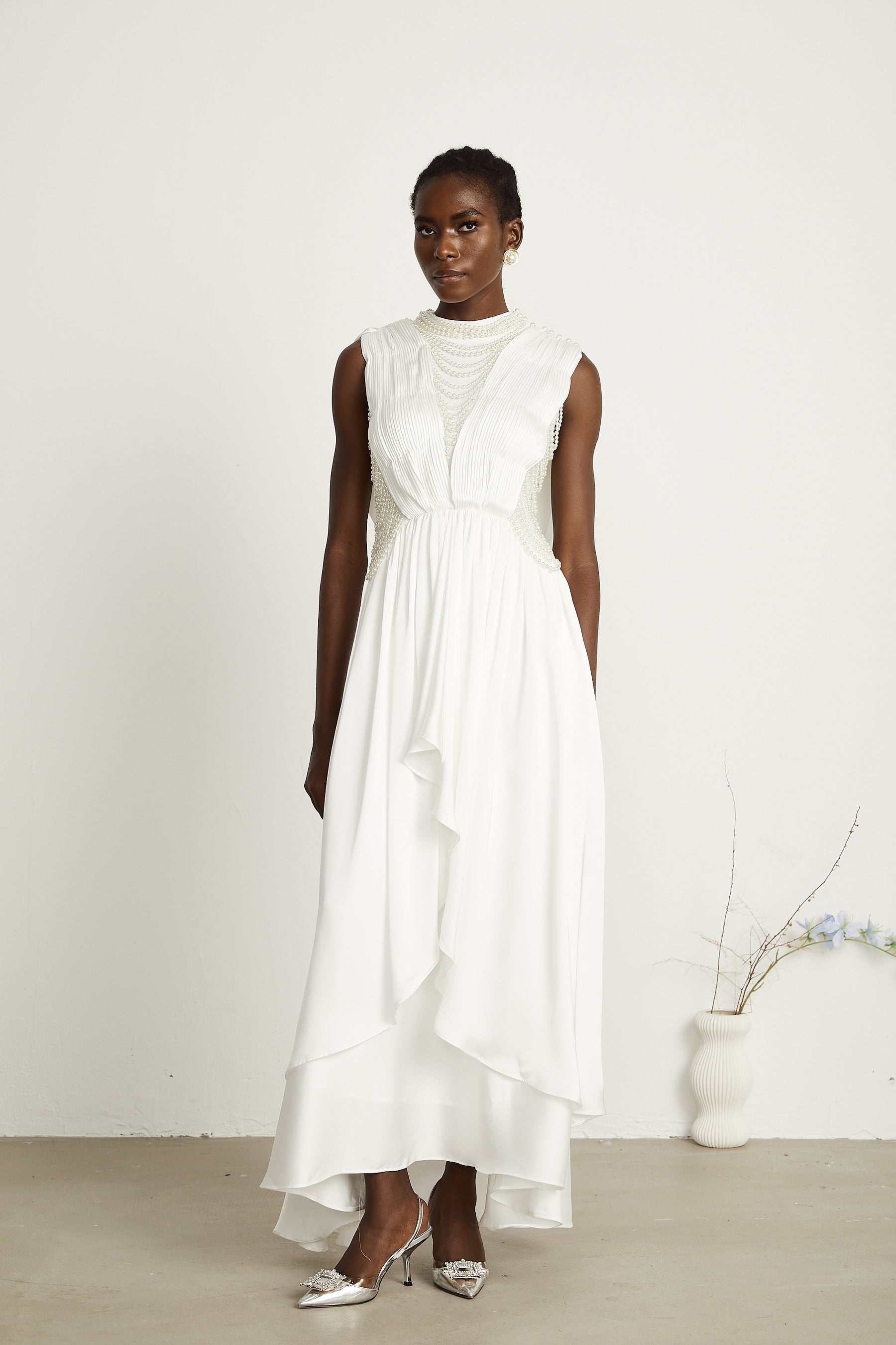 A long white pleated maxi dress with faux pearl embellishments worn by a model standing against a plain background