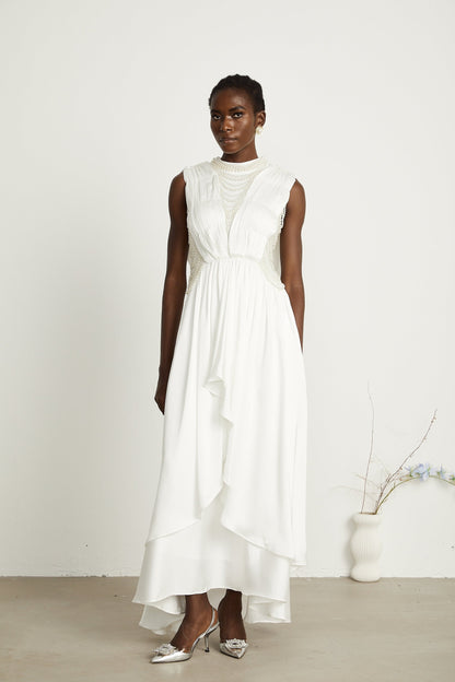 A long white pleated maxi dress with faux pearl embellishments worn by a model standing against a plain background