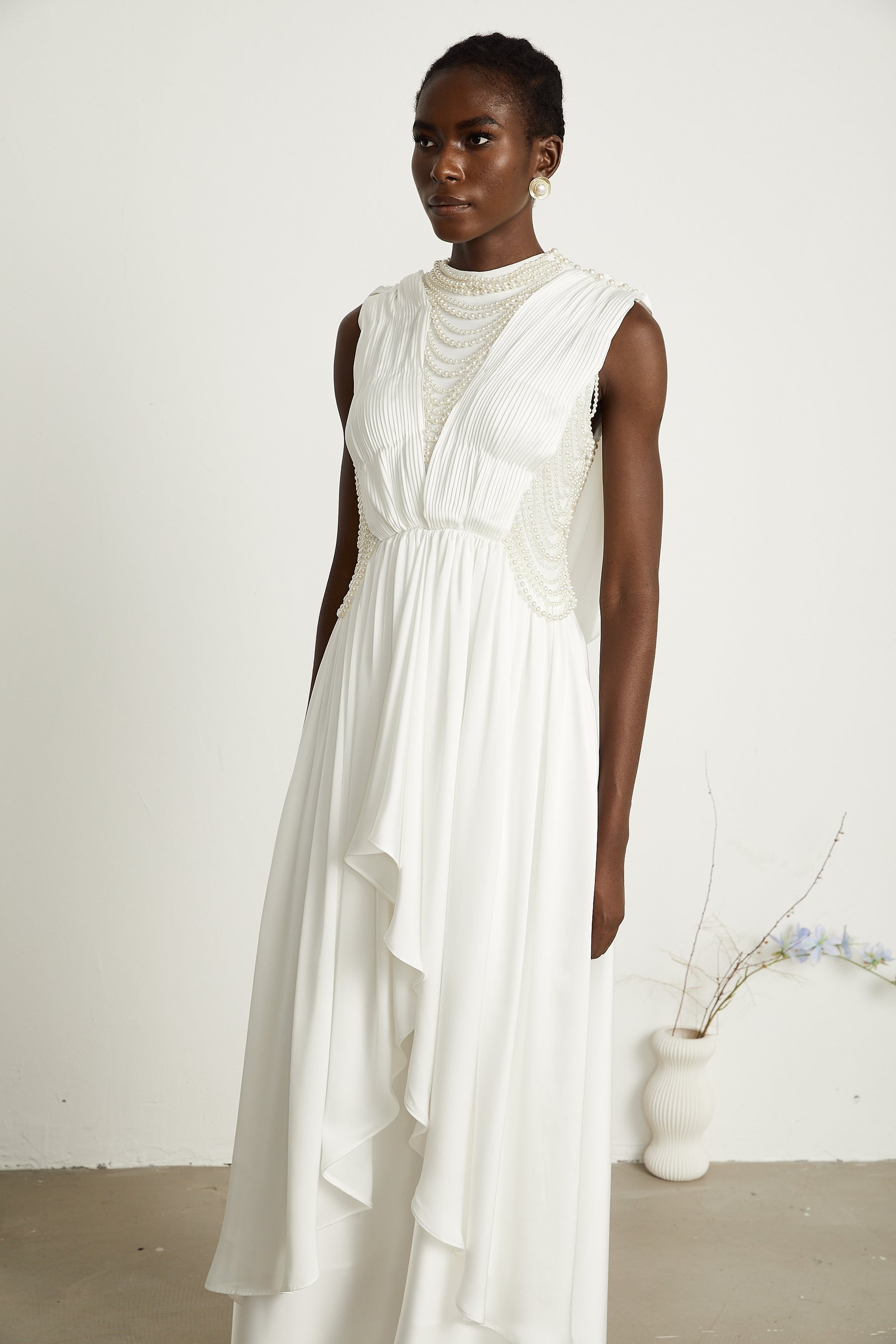 A long white pleated maxi dress with faux pearl embellishments worn by a model standing against a plain background