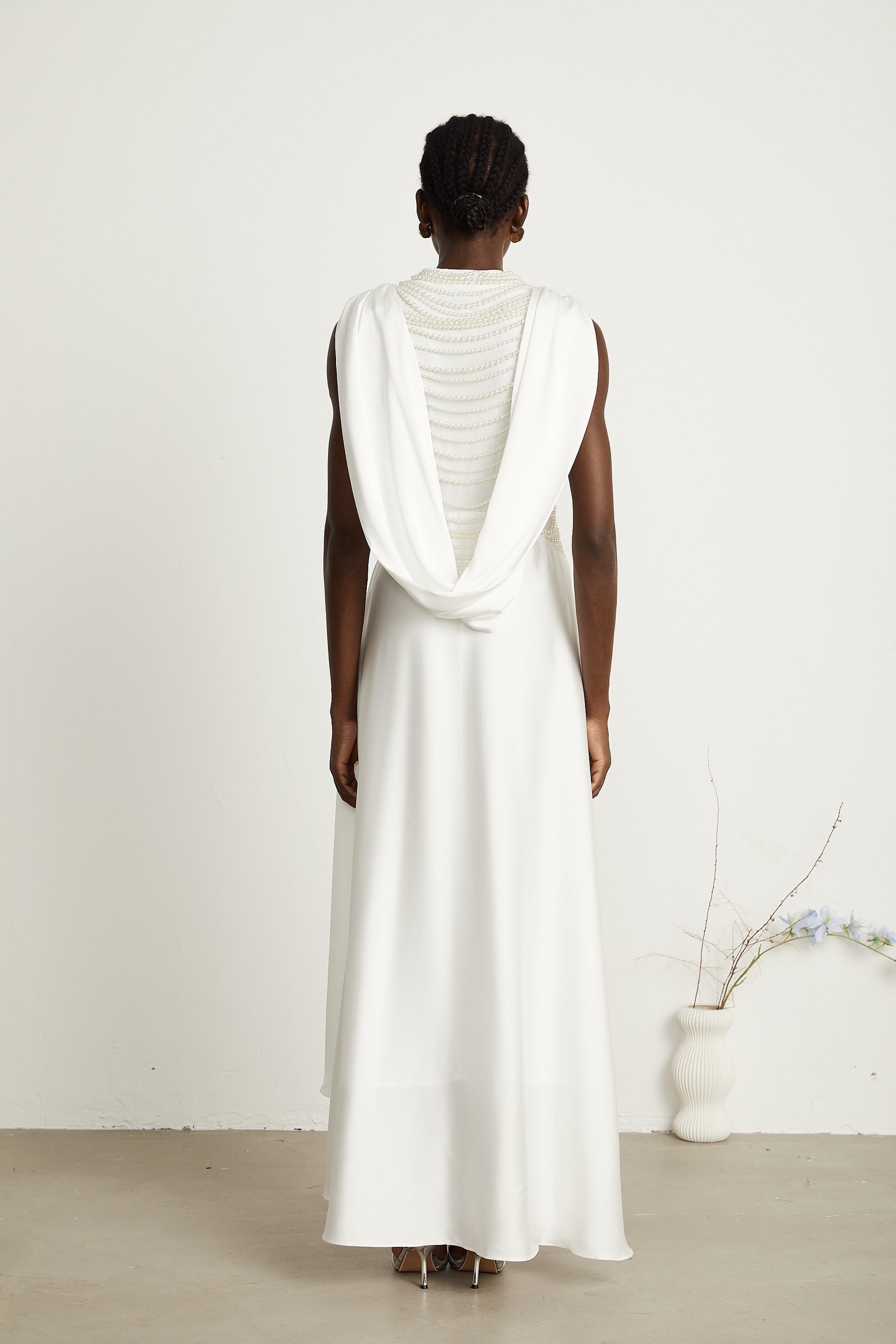 A long white pleated maxi dress with faux pearl embellishments worn by a model standing against a plain background