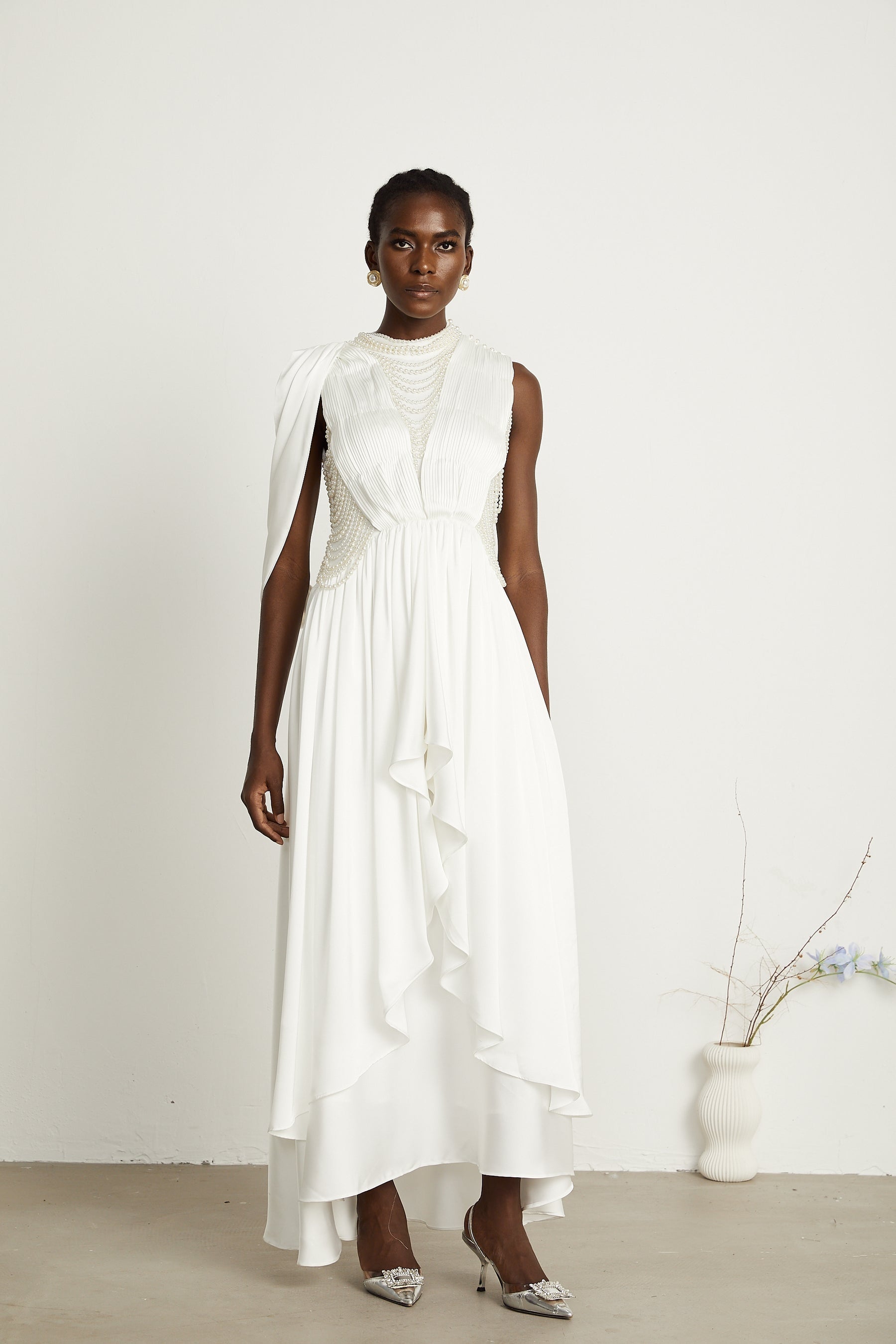 A long white pleated maxi dress with faux pearl embellishments worn by a model standing in a neutral setting