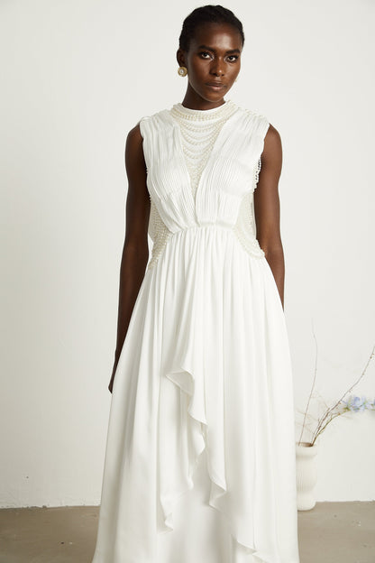 A long white pleated maxi dress with faux pearl embellishments worn by a model standing in an elegant pose