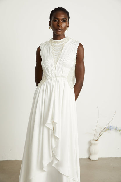 A white floorlength maxi dress with pleated fabric and faux pearl embellishments worn by a model standing in a neutral setting