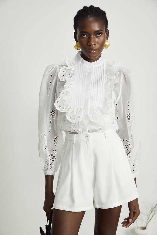 Woman wearing matching white ruffled shirt with cutouts and shorts set standing against plain background