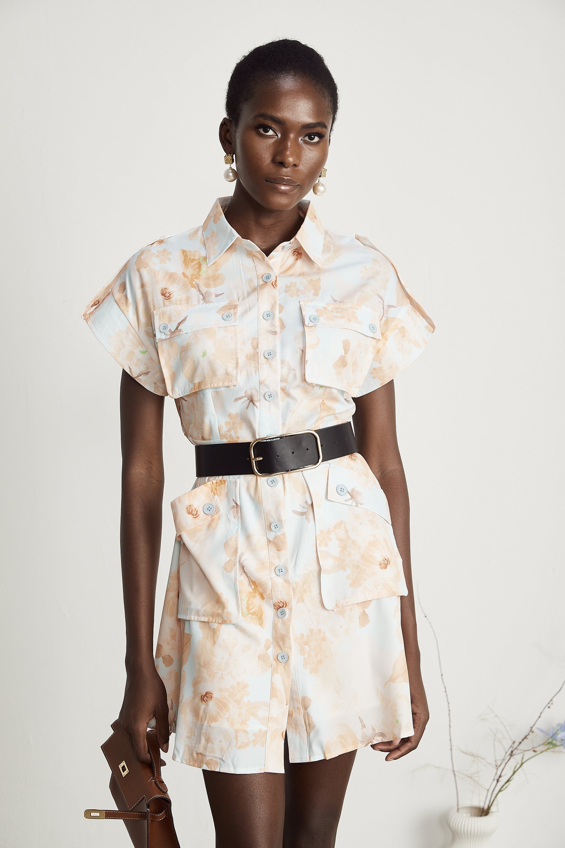 Woman wearing short white belted shirt dress with collar and buttons standing against plain background