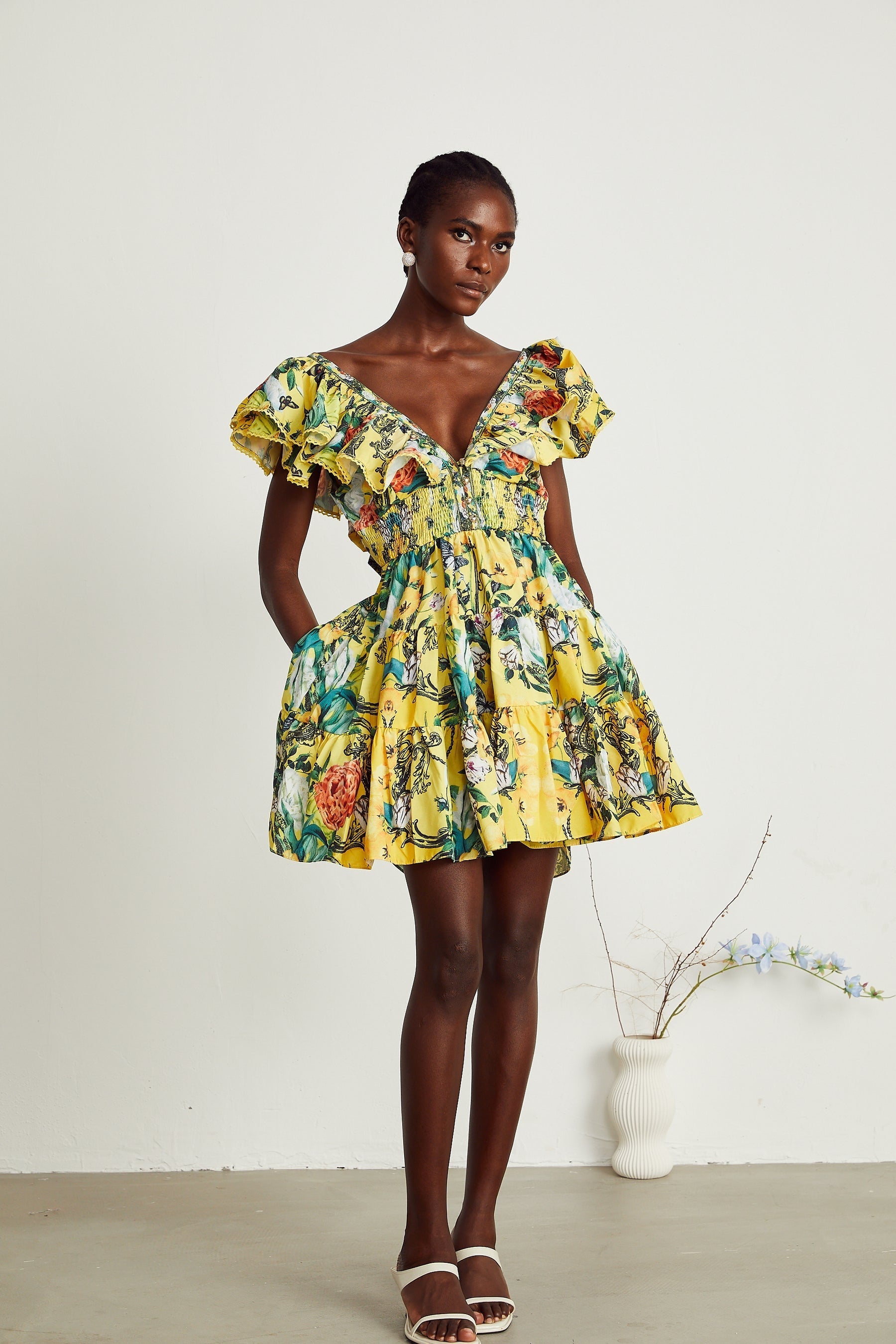 Young woman wearing a yellow mini dress with ruffles and floral print standing against a plain background